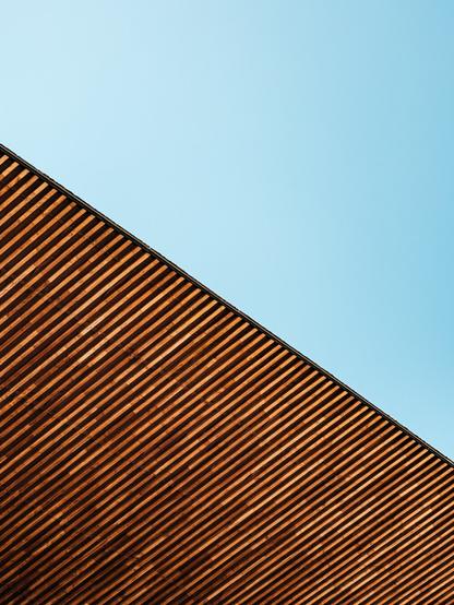 Diagonal view of a wooden slatted roof casting alternating light and dark lines against a clear blue sky. The minimalist composition contrasts the warm texture of the wood with the cool, empty sky.