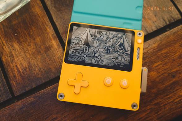 A photo of the Playdate console on a wooden table showing the game Dig Dig Dino on the screen.