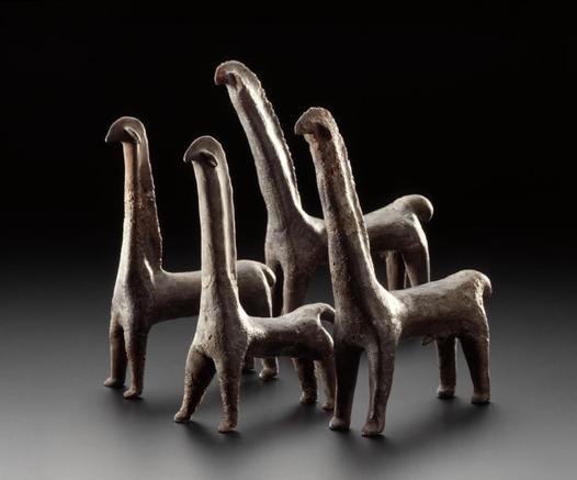 A group of five stylized horse figurines made of clay, arranged on a dark surface with dramatic lighting. The figures have elongated necks, simplified heads, and sturdy legs, with minimal detailing.