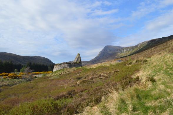A semi-ruined stone tower-like structure against the horizon, one side jutting up higher than the other. The location is a wide open valley of rough grasses and heather, some dark woods off to the left. In the distance, Ben Hope jutting up into a sky that's largely blue, streaked with white cloud.