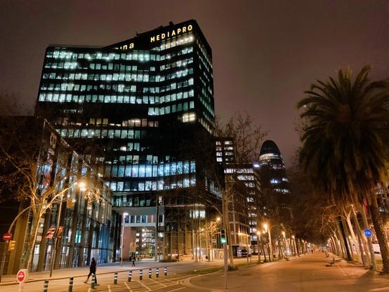 Aquesta imatge nocturna mostra un carrer tranquil de Barcelona, Espanya, amb l’edifici Mediapro il · luminat al fons. L'escena transmet una sensació de calma i quietud, amb pocs vianants visibles. Els arbres alineats al llarg de la vorera i la il·luminació suau creen una atmosfera serena. El modern edifici Mediapro, amb les seves finestres il·luminades, contrasta amb les palmeres i l'atmosfera nocturna més fosca, mostrant un contrast entre tecnologia i natura.
