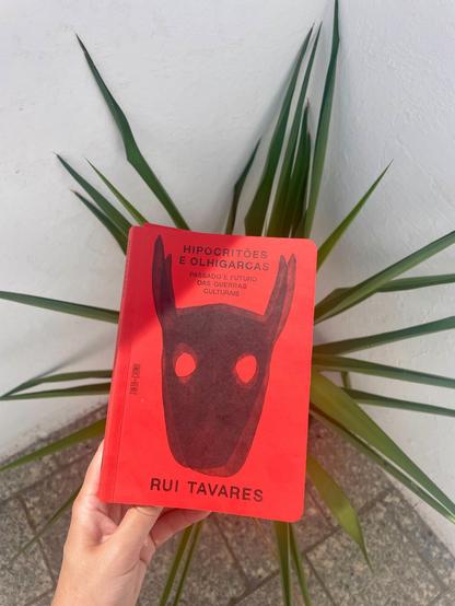 Hand holding a red book about cultural wars by Rui Tavares, a plant in the back