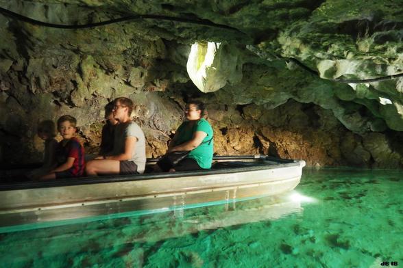 Farbfoto, aufgenommen bei einem Besuch der Wimsener Höhle bei Hayingen auf der Schwäbischen Alb. Diese Wasserhöhle ist im Kahn befahrbar - im türkisgrünen Wasser befindet sich vor der gegenüberliegenden Höhlenwand ein flaches Boot mit vier Besuchern darin.