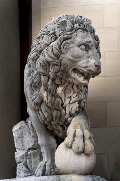 A stone sculpture of a majestic lion, positioned in front of a light-colored wall. The lion is depicted with a fierce expression, its mouth slightly open showing teeth. It has an intricately carved mane with detailed curls and waves. The lion's right paw rests on a smooth stone sphere. The sculpture shows fine craftsmanship in its realistic portrayal of the lion's muscular body and expressive face.