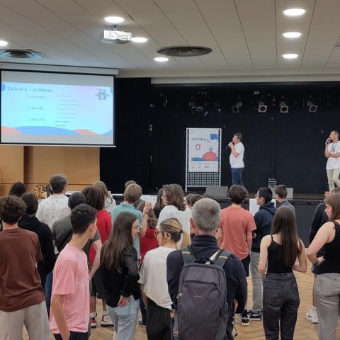 Photo de l'arrière de la salle où l'on voit des jeunes et une estrade avec 2 présentateurs et un diaporama