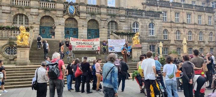 Eine Gruppe von Menschen versammelt sich auf dem Platz vor dem historischen Kasseler Rathaus. Auf der breiten Treppe vor dem Gebäude hängen zwei Banner. Das linke Banner fordert legale Wagenplätze und verweist auf ein Beispiel aus dem Landkreis Kassel. Das rechte Banner trägt die Aufschrift „Wagenleben – gemeinsam solidarisch“ und zeigt eine gezeichnete Szene mit einem Bauwagen. Eine Person spielt Gitarre, während andere zuhören. Im Vordergrund stehen oder sitzen etwa 30 Personen, einige mit Rucksäcken und Fahrrädern. Zwei goldene Löwenfiguren flankieren die Treppe.