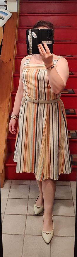 White linen sundress with thin vertical yellow, green and orange stripes. White belt and slingback flats with bows. Gold jewelry.