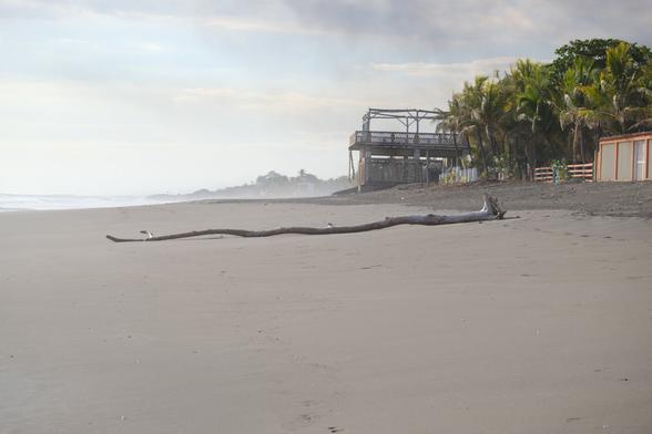 Looking north from Desperados on the beach in Las Peñitas with the roiling surf producing salty mist over the beach. An empty stretch of sand with a large, lone piece of driftwood in the foreground.