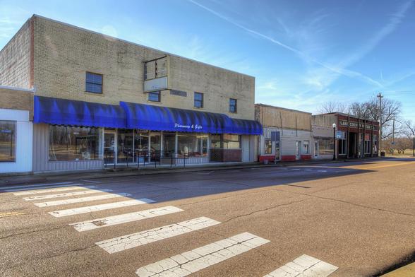 A small town street features a pharmacy and adjacent buildings, with a blue awning and a clear, sunny sky above