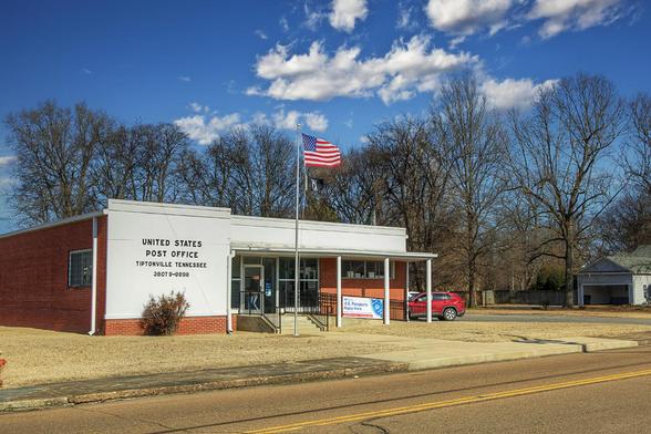 A small United States Post Office building is situated in a rural area, with an American flag flying prominently in front.