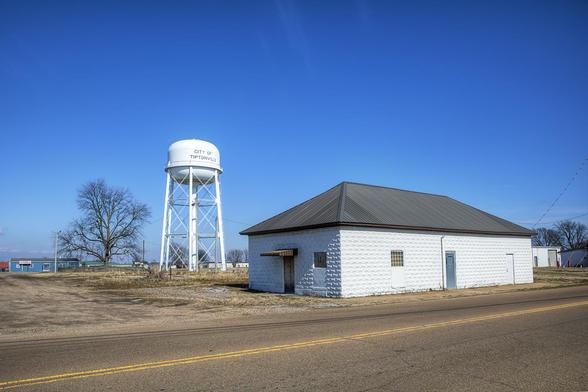 A white water tower marked "City of Tiptonville" rises above a flat landscape near a small building with a metal roof.