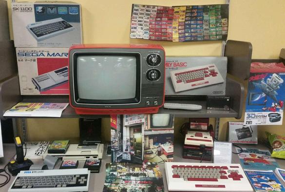 A Sega SG-1000 Mark III and SK-1100 keyboard on the left, and a Famicom with a Disk System and Family Basic and keyboard on the right. Ads, photos, and manuals are scattered about. A lovely orange-red Toshiba Blackstripe sits in the center.
