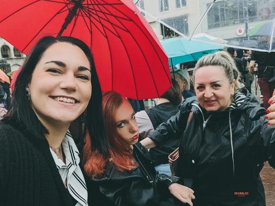 Franzi, Slavi und ich stehen eng beieinander im Regen, auf dem Slutwalk. Links ist Franzi mit schwarzem Haar, Nasenpiercing und gestreifter Bluse. In der Mitte bin ich mit rot gefärbten Haaren und Lederjacke und mit einem ernsten Blick. Rechts ist Slavi mit blondem Dutt und Regenjacke, die einen durchsichtigen Schirm hält. Über der Mitte spannt sich ein leuchtend roter Regenschirm. Im Hintergrund sieht man weitere Menschen mit Regenschirmen auf einer belebten Straße.