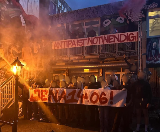 mehrere Menschen stehen hinter einem weißen BAnner auf welchem in rot JENA 14.06.! steht an der Seite eine Eisentreppe, Oben nochmal einige Menschen hinter einem weißen Banner auf welchem "Antifa ist notwendig!" steht