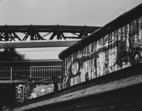 In what appears to be a desolate urban landscape, a moving train glides across a bridge in the background. The scene is defined by stark contrasts and a sense of decay, suggesting that the image was taken in Europe, likely in an aging part of a city.