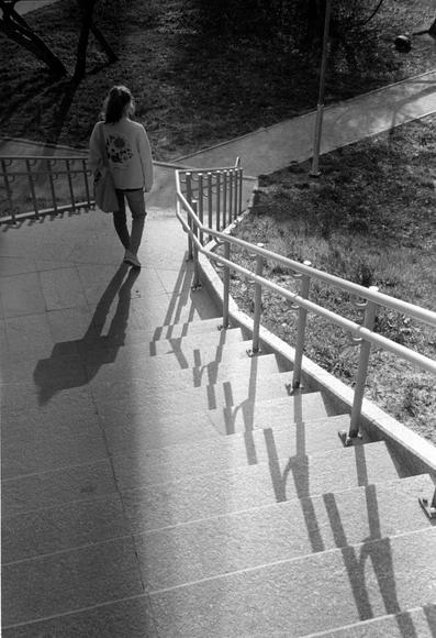Monochrome photograph shows a girl descending a concrete staircase in a park. The sun is bright. Contrasting shadows fall from the girl and from the staircase railing.