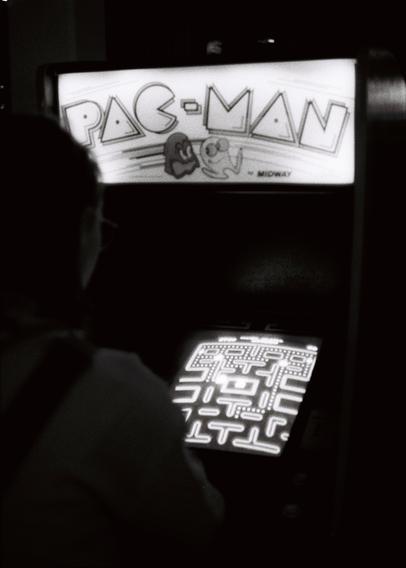 A very dark, black-and-white photograph of a woman playing a Pac-Man arcade game. You know this because a large sign above it says ‘Pac-Man’, and because the good old slightly curved CRT monitor glows with a maze of lines, dots and ghosts.