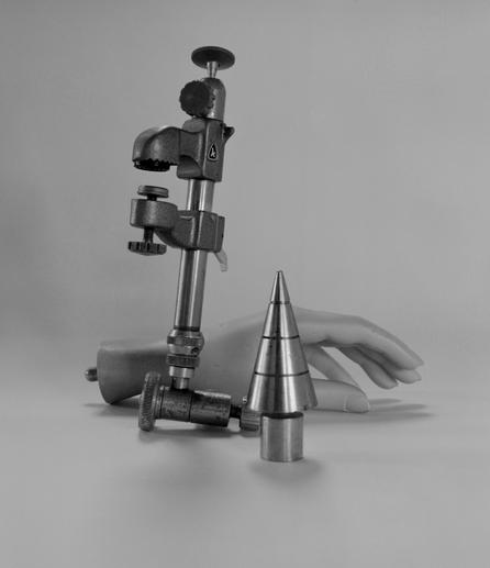 A black and white photograph which shows a mannequins hand towards the back, in the mid-ground is a large, old clamp on it's side and in front is a plumb bob which is stood on it's base, it is conical with rings etched into the metal.