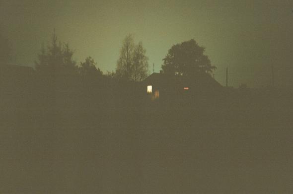 A photo of distant silhouettes of a house and some trees surrounding it against a brown-cyan backdrop. One window has lights on and it is by far the brightest point of the photo