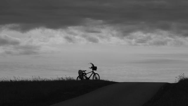 Schwarz weiß. Fahrradsilhouette auf einer Anhöhe. Oben graue Wolkendecke. In der Mitte weiße Wolken mit dem Fahrrad unten dunkler Boden