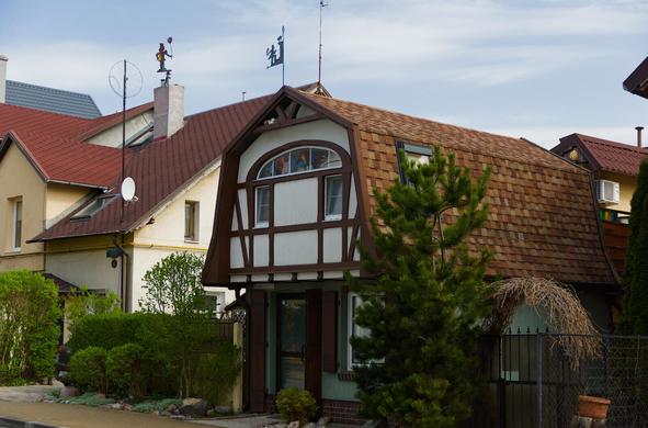 The image captures a quaint, two-story house nestled in what appears to be a residential neighborhood. The architecture is reminiscent of traditional designs with its steeply pitched gable roofs and half-timbered facade painted white, accented by dark brown timbers on the upper story windows and door frame.

The house stands out against a backdrop of other residential buildings that share similar architectural styles but with varying colors like beige and red roofs. The sky above is partly cloudy.

In front of the house, there's a small area with shrubs and trees, adding to the homely charm. A couple of plants are visible on the right side of what seems to be a porch adjacent to the entrance door, which features a classic design with glass panes framed by brown trim.