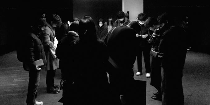 Group of people standing in a circle with heads bowed, in a dimly lit room. Some individuals wear face masks and hold papers or bags. Wooden floor and dark walls enhance the subdued atmosphere.