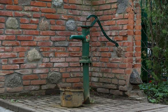 The image captures a green metal hand pump, showing signs of weathering with its peeling paint, stands prominently in the foreground on a brick pavement. Under the pump there is an old plastic bucket.

Behind the pump, there's a red brick wall that adds to the rustic ambiance. Interspersed within this brick structure are several round stones of varying sizes and colors, adding texture and depth to the scene.

To the right side of the photo, greenery peeks into view from behind the bricks, hinting at nature reclaiming its space amidst human-made structures. The overall composition of the image suggests a quiet corner perhaps in an old town or rural setting, where time seems to have stood still.