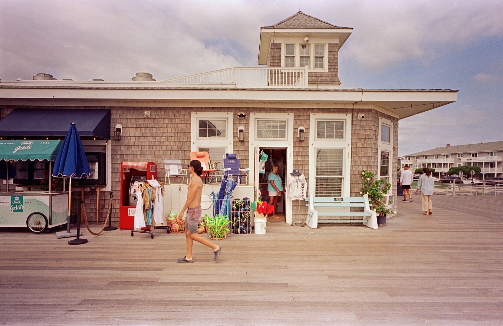 A shirtless man walks down the boardwalk past a cedar shingle covered storefront offering beach chairs, inflatables, and clothing. A gelato stand is visible in front of the next storefront over at left.
