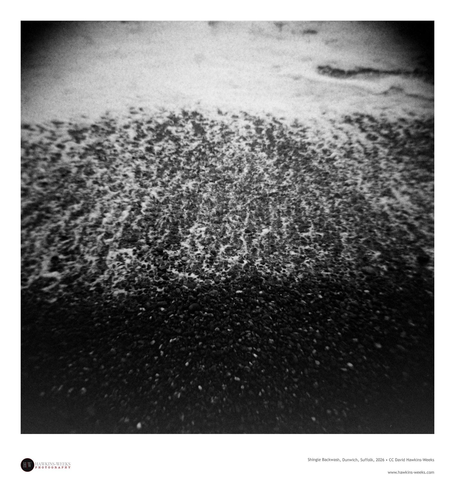 A high-angle, black and white square photograph captures ocean water washing over a pebble beach. The scene is divided into distinct textures and tones, starting with the smooth, light gray surface of the incoming tide at the top. In the middle section, turbulent, foamy white water swirls around tightly packed stones as the wave recedes, while the bottom section is dominated by dark, wet pebbles fading into deep shadow. The image features a strong vignette effect with heavily blurred, darkened edges and a pronounced film grain that creates a moody, lo-fi aesthetic.