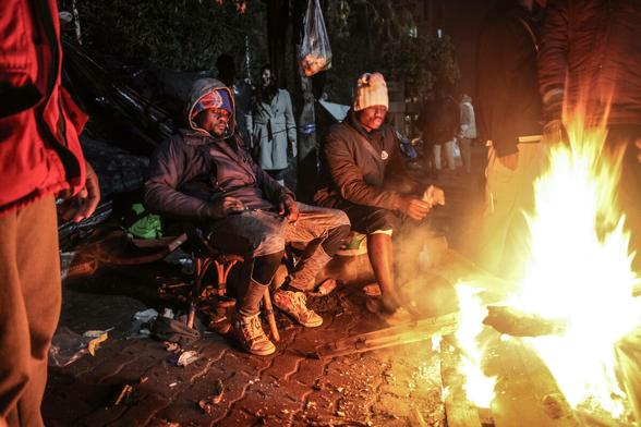 Eine Gruppe ärmlich gekleidter schwarzen Migrant sitzt am Feuer, einer ruht erschöpft