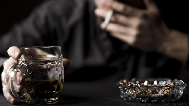 Ein Glas Alkohol und ein voller Aschenbecher, dazu unscharf im Hintergrund ein Mann, der das Glas in der Hand hält und an einer Zigarette zieht.