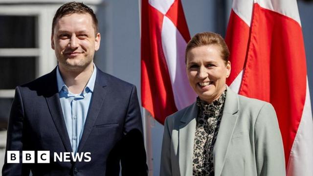 Greenland's Prime Minister Jens-Frederick Nielsen smiles alongside Danish PM Mette Frederiksen, both stood in front of flags outside Frederiksen's residence.