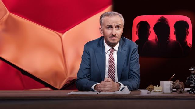 Jan Böhmermann sitzt am Schreibtisch. Rechts neben ihm ist eine Darstellung von drei gesichtslosen Silhouetten vor einem roten Hintergrund abgebildet.