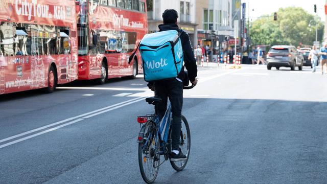 Ein Radfahrer mit einem großen Rucksack auf dem Rücken mit der Aufschrift WOLT fährt auf einer Straße in Köln neben zwei Bussen mit der Aufschrift City Tour 