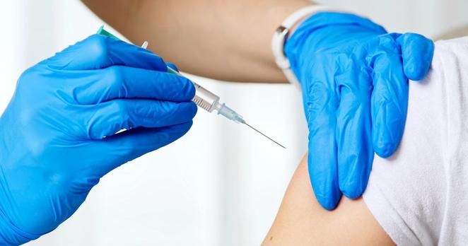 Ein Oberarm, an den eine Spritze angesetzt ist, die mit Händen in blauen Handschuhen gehalten wird.