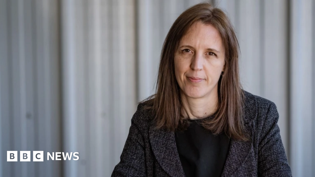 Catherine Braun in a grey suit sat in front of a grey background looking in to the camera 