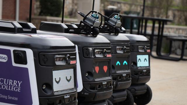 Four delivery robots lined up outdoors, featuring animated digital faces: two eyes with a neutral expression, a heart symbol, and a smiling face with upward eyes. They are branded with 'Curry College' on their sides.