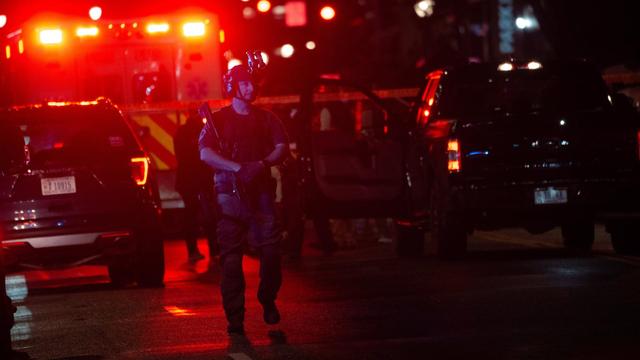 Ein mit Gewehr und Helm ausgestatteter Polizist bewacht einen Tatort im Dunkeln. Im Hintergrund sind die Lichter eines Krankenwagens zu sehen. 