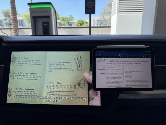 The interior dash of a Rivian. There's a large built-in touchscreen on the left and an iPad mini mounted on the right. The large screen shows a person holding a menu printed in Japanese, the iPad has GoodNotes and a knitting pattern.