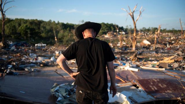 Ein Mann mit schwarzem Cowboyhut steht mit gesenktem Kopf vor den Überresten eines durch Tornados zerstörten Gebäudes.