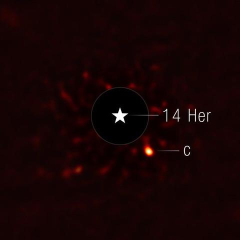 This image shows the exoplanet 14 Herculis c. The view is mostly black, with very faint red splotches in the central region of the image. At the center of the image, there is a black circle, and in the center of that, there is a star symbol representing a real star. This black circle blocks the light from the host star. To the lower right of the circle is a fuzzy bright orange circle, which is the exoplanet.