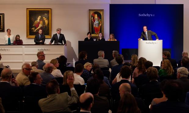 A scene from an auction: a man stands at a rostrum next to several people and in front of a crowd.