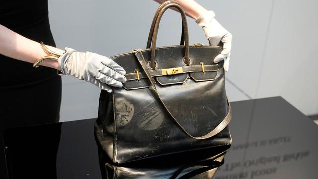 Zu sehen sind zwei Hände in weißen Handschuhen, die eine Designer-Handtasche auf einem Podest präsentieren.