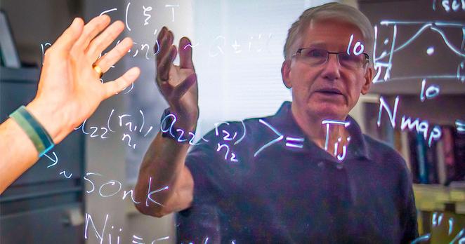 A man with white hair and glasses is seen reflected in a shiny blackboard.