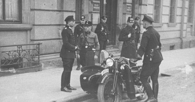 Uniformierte Männer mit einem Motorrad vor einem Gebäude aus dem 19. Jahrhundert.