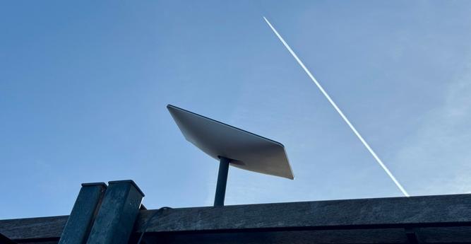 A starlink dish against a bright sky as a jet flies past overhead.