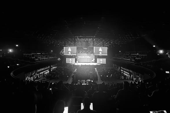 A black and white photograph of a large concert venue filled with audience members, featuring a stage with large video screens displaying "Taylor Otwell" and a man standing at a podium.