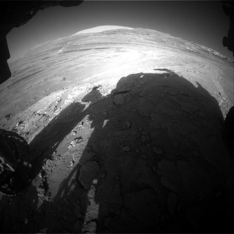 An extreme wide-angle, grayscale photo from the Mars surface shows uneven terrain ahead of the rover, covered in numerous small to medium-sized rocks, all light gray and illuminated by sunlight. Impressions in the middle ground ahead form swooping backward Cs, and in the far distance a mountain rises from the ground, but looks very broad and squat, because of the distortion from the wide-angle lens. Parts of the rover are visible around the sides and bottom of the image, and its shadow fills much of the foreground.