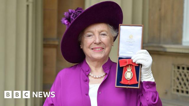Dame Stephanie Shirley wearing a purple hat, matching purple dress, and a pearl necklace, smiles at the camera while holding a medal outside Buckingham Palace after she was made a member of the Order of the Companions of Honour