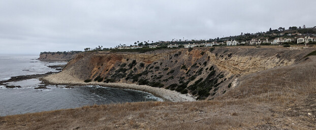 Looking across a cove with a narrow rocky beach from the top of the coastal bluffs on a gloomy day. Layers of stone curve upward in the middle, and bushes clump on the dirt escarpment. Houses line the hill in the distance.